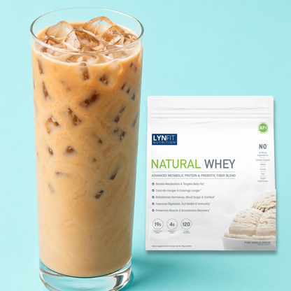 A tall glass of iced coffee with creamy brown liquid and ice cubes stands beside a white bag of Lynfit Nutritionals Natural Whey vanilla flavor, set against a light blue background.